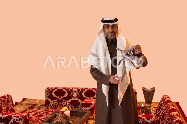 جلسة برية في الصحراء السعودية، أساليب عريقة في كرم الضيافة، النظر إلى الكاميرا بإيماءات السرور، حسن استقبال وإكرام الضيوف، بورتريه مقرب لسيد عربي سعودي خليجي مبتسم يرتدي الغترة والثوب التقليدي يقوم بصب القهوة العربية، خلفية بيج.