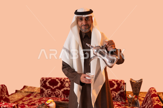 عادات وتقاليد سعودية في جلسات شعبية، أساليب عريقة في كرم الضيافة، النظر إلى الكاميرا بإيماءات السرور، حسن استقبال وإكرام الضيوف، بورتريه مقرب لسيد عربي سعودي خليجي مبتسم يرتدي الغترة والثوب التقليدي يقوم بصب القهوة العربية، خلفية بيج.