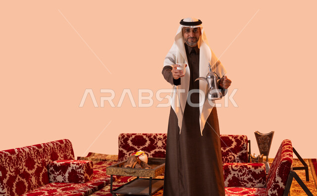 جلسة شعبية في الصحراء السعودية، حسن استقبال وإكرام الضيوف، عادات وتقاليد أصيلة في كرم الضيافة، بورتريه لسيد عربي سعودي خليجي مبتسم يرتدي الغترة والثوب التقليدي يقوم بتقديم القهوة العربية بإيماءات السرور والترحيب، منتج محلي وطني، خلفية بيج.