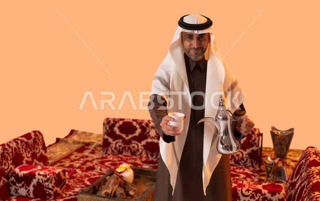 أساليب عريقة في كرم الضيافة، النظر إلى الكاميرا بإيماءات السرور، جلسة شعبية في الصحراء السعودية، حسن استقبال وإكرام الضيوف، بورتريه مقرب لسيد عربي سعودي خليجي مبتسم يرتدي الغترة والثوب التقليدي يقوم بتقديم القهوة العربية، خلفية بيج.