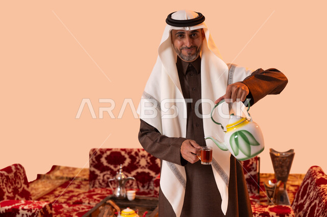 كشتة بر وتخييم شعبية سعودية، النظر إلى الكاميرا بإيماءات السرور والسعادة، الاستمتاع بتناول المشروبات والأعشاب الساخنة، بورتريه مقرب لسيد عربي سعودي خليجي مبتسم يرتدي الزي التقليدي يقوم بصب الشاي، خلفية بيج.