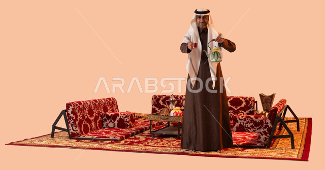 جلسة شعبية في البراري والصحاري السعودية، استقبال وإكرام الضيوف، النظر إلى الكاميرا بإيماءات السرور والسعادة، الاستمتاع بتناول المشروبات الساخنة، بورتريه مقرب لسيد عربي سعودي خليجي مبتسم يرتدي الزي التقليدي يقوم بتقديم الشاي، صورة لكامل الجسم، خلفية بيج.