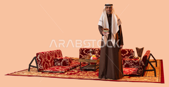 طبق زجاجي مليء بالتمور، حسن الاستقبال وكرم الضيافة,عادات وتقاليد سعودية في جلسات شعبية، محاصيل زراعية وطنية محلية، بورتريه لرجل عربي سعودي خليجي كبير بالسن يرتدي الغترة والثوب التقليدي يحمل بيده تمريه مزخرفة في كشتة برية، صورة الجسم بالكامل،