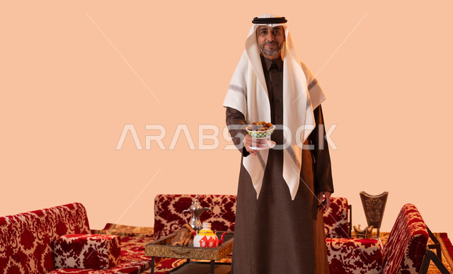 حسن الاستقبال وكرم الضيافة,عادات وتقاليد سعودية في جلسات شعبية، محاصيل زراعية وطنية محلية، طبق زجاجي مليء بالتمور، بورتريه مقرب لرجل عربي سعودي خليجي كبير بالسن يرتدي الغترة والثوب التقليدي يحمل بيده تمريه مزخرفة في كشتة برية، خلفية بيج