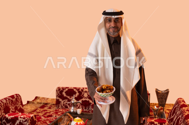 Popular Saudi customs and traditions in honoring the guest by presenting national agricultural products, Bedouin wild sessions, local Saudi crops, a portrait of an old Saudi Arabian Gulf man wearing traditional clothing holding a golden date in his hand, the hospitality of the Saudis, a beige background