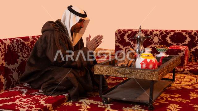 الأجواء الهادئة في المخيمات الشعبيه ،إيماءات التدفئة على الموقد, أسلوب الحياة البدوية القديمة, جلسة بر شبابية شعبية، بورتريه لرجل كبير السن عربي خليجي سعودي يرتدي الزي التقليدي والملابس الشتوية الدافئة يجلس أمام شعلة من النار ،خلفية بيج