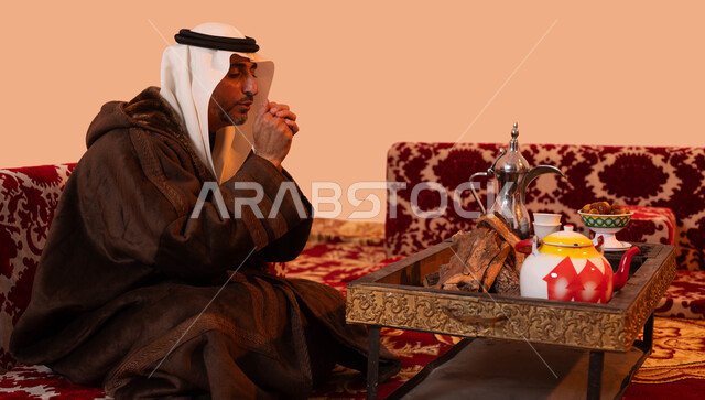 جلسات بر خارجية، النفخ داخل الكفين للشعور بالدفئ، الأجواء الهادئة في المخيمات الشعبيه ،إيماءات التدفئة على الموقد, بورتريه مقرب لرجل كبير السن عربي خليجي سعودي يرتدي الزي التقليدي والملابس الشتوية الدافئة يجلس أمام شعلة من النار ، خلفية بيج