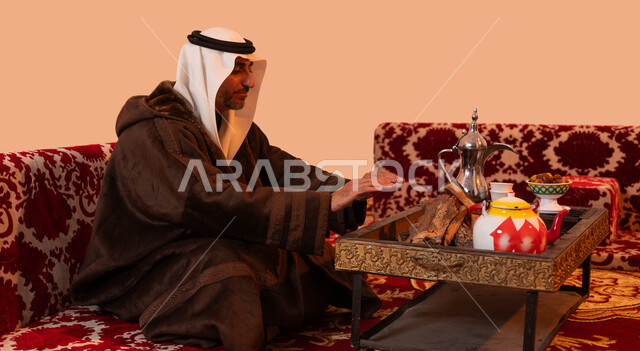 إيماءات التدفئة على الموقد, الأجواء الهادئة في أماكن التخييم، جلسة بر شبابية شعبية، أسلوب الحياة القديمة،  بورتريه مقرب لرجل كبير السن عربي خليجي سعودي يرتدي الزي التقليدي والملابس الشتوية الدافئة يجلس أمام شعلة من النار ، خلفية بيج.