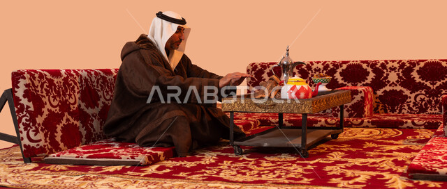 أسلوب الحياة القديمة، الأجواء الهادئة في أماكن التخييم، جلسة بر شبابية شعبية، إيماءات التدفئة على الموقد,  بورتريه لرجل كبير السن عربي خليجي سعودي يرتدي الزي التقليدي والملابس الشتوية الدافئة يجلس أمام شعلة من النار ، خلفية بيج.