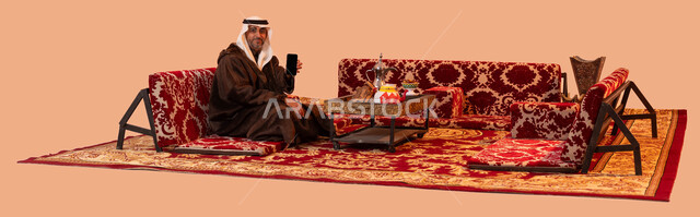 إيماءات البرد والسعادة بالأجواء الشتوية، عرض شاشه سوداء فارغة، جلسات بر شعبيه بدوية، استخدام الجوال في التسوق الالكتروني، بورتريه لرجل كبير السن عربي خليجي سعودي مبتسم يرتدي الملابس الدافئة الشتوية والزي التقليدي يحمل بيده الهاتف المحمول ،خلفية بيج.