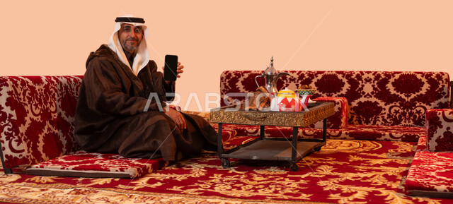 عرض شاشه سوداء فارغة، جلسات بر شعبيه بدوية، استخدام الجوال في التسوق الالكتروني، بورتريه لرجل كبير السن عربي خليجي سعودي مبتسم يرتدي الملابس الدافئة الشتوية والزي التقليدي يحمل بيده الهاتف المحمول ،إيماءات البرد والسعادة بالأجواء الشتوية، خلفية بيج.
