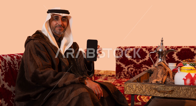 جلسات بر شعبيه بدوية، استخدام الجوال في التسوق الالكتروني، بورتريه مقرب لرجل مسن عربي خليجي سعودي مبتسم يرتدي الملابس الدافئة الشتوية والزي التقليدي يحمل بيده الهاتف المحمول بشاشه سوداء فارغة، إيماءات البرد والسعادة بالأجواء الشتوية، خلفية بيج.