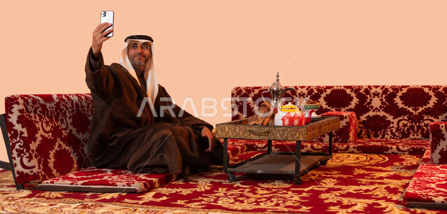 أجواء التخييم الهادئة في فصل الشتاء، توثيق اللحظات السعيدة في جلسات البر الشعبية بالتقاط صور تذكارية عبر الهاتف المحمول، بورتريه لرجل كبير السن عربي سعودي خليجي يرتدي اللباس الشتوي الدافئ والزي التقليدي يحمل بيده الجوال، خلفية بيج