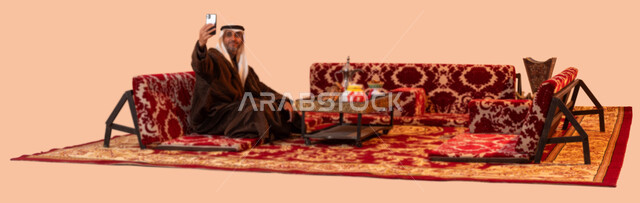 توثيق اللحظات السعيدة في جلسات البر الشعبية بالتقاط صور تذكارية عبر الهاتف المحمول، بورتريه لرجل كبير السن عربي سعودي خليجي يرتدي اللباس الشتوي الدافئ والزي التقليدي يحمل بيده الجوال، أجواء التخييم الهادئة في فصل الشتاء، خلفية بيج