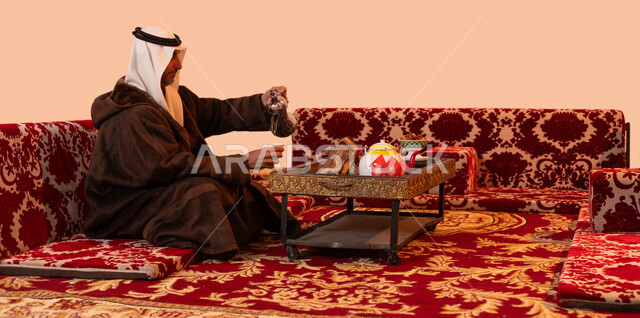 جلسات بدويه شعبيه ،نزهه بريه في الصحراء، عادات وتقاليد سعودية اصيلة في تقديم القهوة العربية، مفهوم حسن الضيافه والكرم، بورتريه لرجل مسن عربي خليجي سعودي مبتسم يرتدي الزي التقليدي والملابس الشتوية الدافئة يقوم بصب القهوة باستخدام الدلة النحاسية، خلفية بيج
