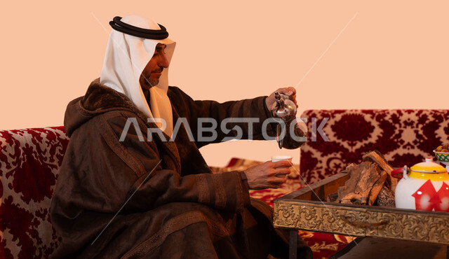 جلسات شعبية خارجية، عادات وتقاليد سعودية اصيلة في تقديم القهوة العربية، مفهوم حسن الضيافه والكرم، بورتريه مقرب لرجل مسن عربي خليجي سعودي مبتسم يرتدي الزي التقليدي والملابس الشتوية الدافئة يقوم بصب القهوة باستخدام الدلة النحاسية، خلفية بيج