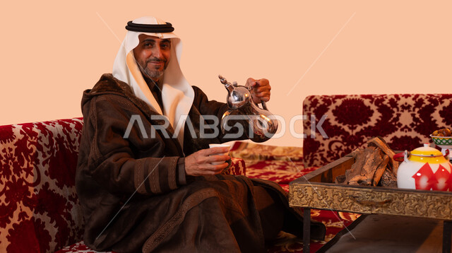 النظر إلى الكاميرا، جلسات شعبية خارجية، عادات وتقاليد سعودية اصيلة في تقديم القهوة العربية، مفهوم حسن الضيافه والكرم، بورتريه مقرب لرجل مسن عربي خليجي سعودي مبتسم يرتدي الزي التقليدي والملابس الشتوية الدافئة يقوم بصب القهوة باستخدام الدلة النحاسية، خلفية بيج