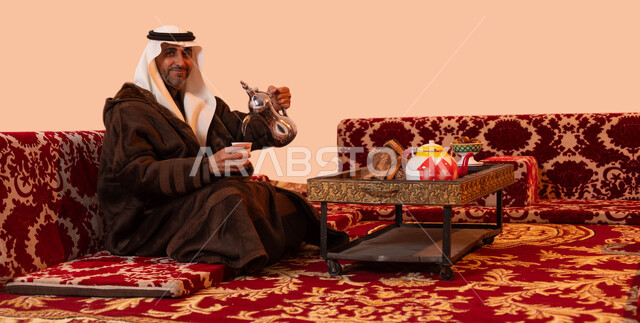 جلسات شعبية خارجية شبابية، عادات وتقاليد سعودية اصيلة في تقديم القهوة العربية، مفهوم حسن الضيافه والكرم، بورتريه لرجل كبير السن عربي خليجي سعودي مبتسم يرتدي الزي التقليدي والملابس الشتوية الدافئة يقوم بصب القهوة باستخدام الدلة النحاسية، خلفية بيج.