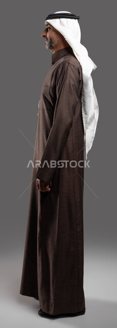 الوقوف بثقة بالنفس واستقامة، النظر إلى شيء ما، بورتريه مقرب من الجانب لرجل كبير السن عربي خليجي سعودي مبتسم يرتدي الزي التقليدي والشماغ يقف بثقة واستقامة ،مفهوم الاناقة والاهتمام بالمظهر الخارجي، خلفية رمادية