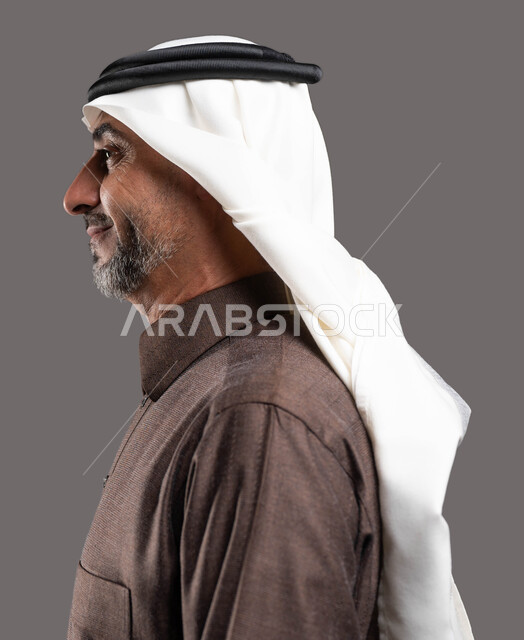 الوقوف بثقة واستقامة، بورتريه مقرب من الجانب لرجل كبير السن عربي خليجي سعودي مبتسم يرتدي الزي التقليدي والشماغ ينظر إلى شيء ما، مفهوم الاناقة والاهتمام بالمظهر الخارجي، خلفية رمادية