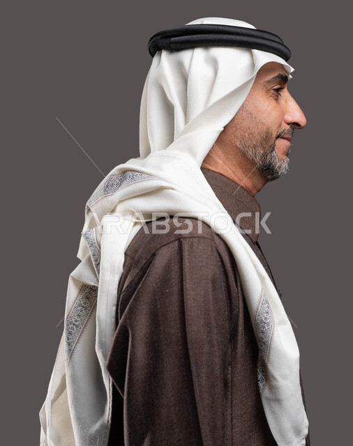 مفهوم الاناقة والاهتمام بالمظهر الخارجي، الوقوف بثقة واستقامة، بورتريه مقرب من الجانب لرجل كبير السن عربي خليجي سعودي مبتسم يرتدي الزي التقليدي والشماغ ينظر إلى شيء ما، خلفية رمادية