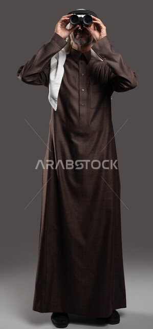 التخطيط المستقبلي للطموحات والأهداف، النظر من المنظار ثنائي العينين لرؤية الاشياء البعيدة، بورتريه لرجل كبير السن عربي خليجي سعودي يرتدي الثوب التقليدي والشماغ يقوم بمشاهدة أحداث معينة عبر التلسكوب، صورة الجسم بالكامل، خلفية رمادية