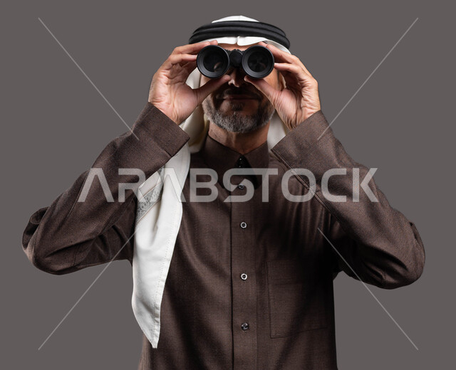 مشاهدة أحداث معينة عبر التلسكوب، النظر من المنظار ثنائي العينين لرؤية الاشياء البعيدة، بورتريه مقرب لرجل كبير السن عربي خليجي سعودي يرتدي الثوب التقليدي والشماغ يقوم بالتخطيط المستقبلي للأهداف و الطموحات، خلفية رمادية