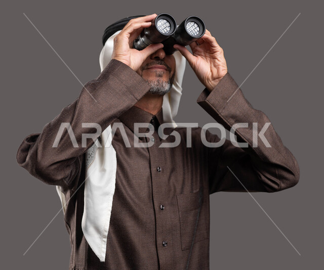 النظر من المنظار ثنائي العينين لرؤية الاشياء البعيدة ، حب الاستكشاف والمغامرة، بورتريه مقرب لرجل كبير السن عربي خليجي سعودي يرتدي الثوب التقليدي والشماغ يحمل بيده التلسكوب و يقوم بالتخطيط المستقبلي للأهداف و الطموحات، خلفية رمادية
