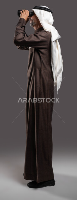 حب الاستكشاف والمغامرة، رؤية الاشياء البعيدة عبر التلسكوب، بورتريه من الجانب لرجل مسن عربي خليجي سعودي يرتدي الثوب التقليدي والشماغ ينظر من خلال منظار الرؤية, التخطيط المستقبلي للأهداف و الطموحات، صورة الجسم بالطول الكامل، خلفية رمادية