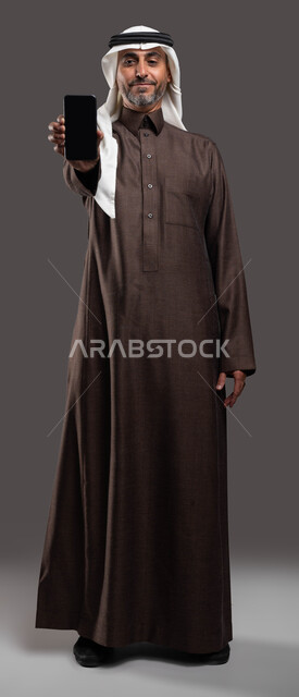 ايماءات الاعجاب باستخدام التقنيه والتكنولوجيا المتقدمة، استخدام الأجهزة الحديثة في تطوير المهارات الشخصية، بورتريه مقرب لرجل كبير السن عربي خليجي سعودي مبتسم يرتدي الزي التقليدي يحمل بيده الجوال بشاشة سوداء فارغة، صورة الجسم بالكامل، خلفية رمادية