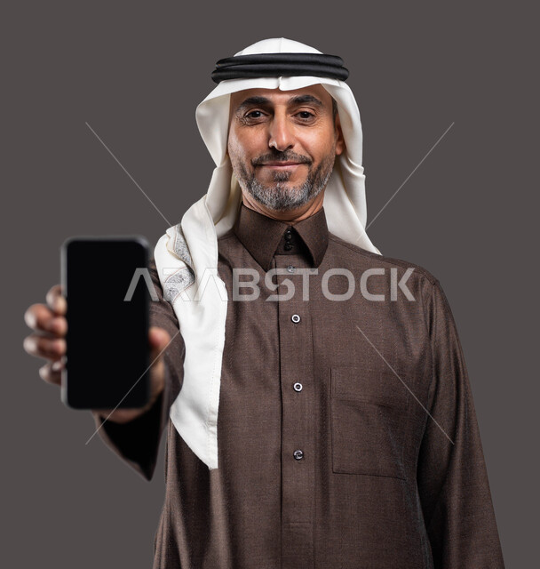 Using modern devices to develop personal skills, gestures of admiration using technology and advanced technology, a close-up portrait of an elderly smiling Saudi Arabian Gulf man wearing traditional clothing holding a mobile phone in his hand with a blank black screen, gray background