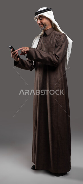 الدردشه والمحادثات الوديه مع الأصدقاء عبر الانترنت, بورتريه لرجل مسن عربي سعودي خليجي مبتسم يرتدي الثوب والشماغ التقليدي يقوم بتصفح مواقع التواصل الاجتماعي عبر الهاتف المحمول , استخدام التقنيات الحديثة المتطورة, صورة الجسم بالطول الكامل, خلفيه رماديه