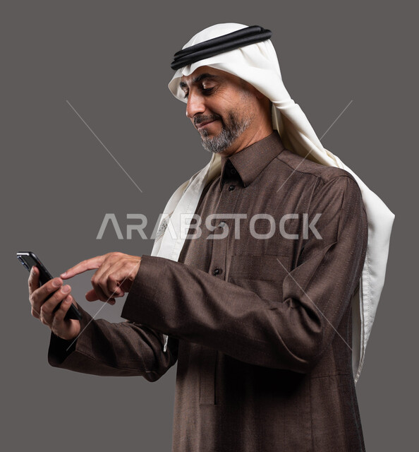 النقر والضغط باستخدام حاسة اللمس، الدردشه والمحادثات الوديه مع الأصدقاء عبر الانترنت, بورتريه لرجل كبير السن عربي سعودي خليجي مبتسم يرتدي الثوب والشماغ التقليدي يقوم بتصفح مواقع التواصل الاجتماعي عبر الهاتف المحمول بإيماءات التركيز, خلفيه رماديه
