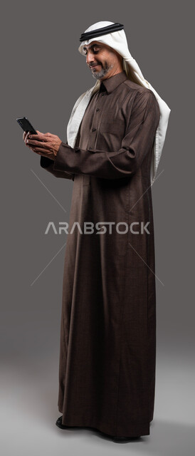 التسويق الالكتروني عبر الانترنت, بورتريه لرجل كبير السن عربي سعودي خليجي مبتسم يرتدي الثوب والشماغ التقليدي يقوم بتصفح مواقع التواصل الاجتماعي عبر الهاتف المحمول بإيماءات التركيز, استخدام التقنيات الحديثة المتطورة, صورة الجسم بالكامل، خلفيه رماديه