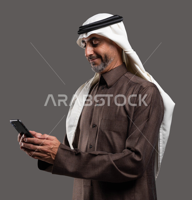 استخدام التقنيات الحديثة المتطورة, الدردشات والمحادثات الوديه مع الأصدقاء عبر الانترنت, بورتريه لرجل كبير السن عربي سعودي خليجي مبتسم يرتدي الثوب والشماغ التقليدي يقوم بتصفح مواقع التواصل الاجتماعي عبر الهاتف المحمول بإيماءات التركيز,  خلفيه رماديه
