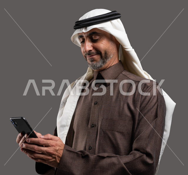 الدردشه والمحادثات الوديه مع الأصدقاء عبر الانترنت, بورتريه لرجل كبير السن عربي سعودي خليجي مبتسم يرتدي الثوب والشماغ التقليدي يقوم بتصفح مواقع التواصل الاجتماعي عبر الهاتف المحمول بإيماءات التركيز, استخدام التقنيات الحديثة المتطورة, خلفيه رماديه
