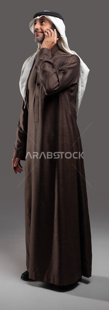 التواصل الودي مع الأصدقاء عبر الانترنت, بورتريه لرجل شايب عربي سعودي خليجي مبتسم يرتدي الثوب والشماغ التقليدي يقوم بإجراء مكالمة  صوتية عبر الهاتف المحمول, استخدام التقنيات الحديثة المتطورة, صورة الجسم بالطول الكامل، خلفيه رماديه