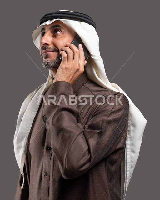 المحادثة والدردشة الوديه مع الأصدقاء عبر الانترنت, بورتريه لرجل شايب عربي سعودي خليجي مبتسم يرتدي الثوب والشماغ التقليدي يقوم بإجراء مكالمة  صوتية عبر الهاتف المحمول, استخدام التقنيات الحديثة المتطورة, خلفيه رماديه
