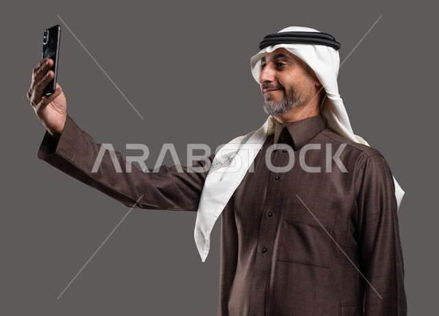 إجراء مكالمات الفيديو مع الأصدقاء والأهل، استخدام التقنيات الحديثة, بورتريه لرجل كبير السن عربي سعودي خليجي مبتسم يرتدي الثوب والشماغ التقليدي يقوم بالتقاط صور السيلفي التذكارية عبر الهاتف المحمول, خلفيه رماديه
