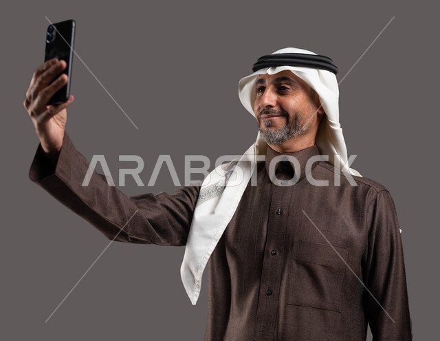 التقاط صور السيلفي التذكارية، الدردشه والمحادثات الوديه مع الأصدقاء عبر الانترنت, بورتريه لرجل كبير السن عربي سعودي خليجي مبتسم يرتدي الثوب والشماغ التقليدي يقوم بإجراء مكالمة فيديو عبر الهاتف المحمول, استخدام التقنيات الحديثة, خلفيه رماديه