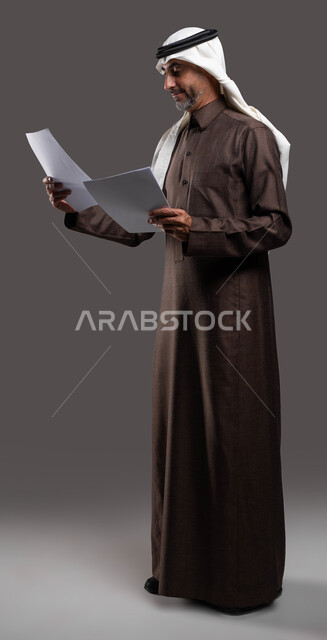 اعداد التقارير الخاصة بالعمل، التدقيق في قراءة الملاحظات، بورتريه لرجل شايب عربي خليجي سعودي مبتسم يرتدي الثوب التقليدي والشماغ يحمل بيده مجموعه من الاوراق البيضاء، مراجعة ومتابعة الخطط والمشاريع المكتبية، صورة الجسم بالطول الكامل، خلفية رمادية .