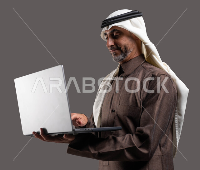 استخدام الحاسوب المحمول لإنجاز المهام، رجل كبير السن عربي سعودي خليجي مبتسم يرتدي الزي السعودي يحمل جهاز اللابتوب، تطور التكنولوجيا والتقنيات الحديثة، إجتماع عمل عن بعد، إجراء مكالمات فيديو عبر جهاز الحاسوب, خلفيه رمادية .