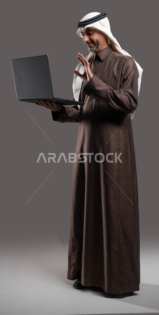 إجتماع عمل عن بعد، إجراء مكالمات فيديو عبر الحاسوب المحمول, إيماءات السلام والتلويح، رجل كبير السن عربي سعودي خليجي مبتسم يرتدي الزي السعودي يحمل جهاز اللابتوب، تطور التكنولوجيا والتقنيات الحديثة، صورة الجسم بالطول الكامل، خلفيه رمادية .