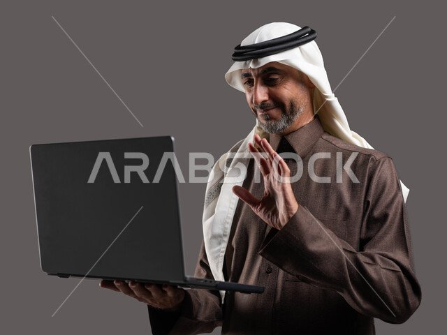 إيماءات السلام والتلويح، رجل كبير السن عربي سعودي خليجي مبتسم يرتدي الزي السعودي يحمل جهاز اللابتوب، تطور التكنولوجيا والتقنيات الحديثة، إجتماع عمل عن بعد، إجراء مكالمات فيديو عبر الحاسوب المحمول, خلفيه رمادية .