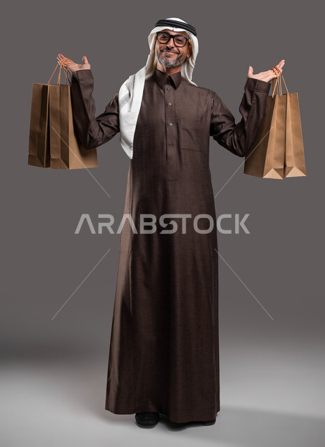 الاستفادة من الحسومات الموسمية، بورتريه لرجل مسن عربي خليجي سعودي يرتدي الثوب التقليدي والغترة والنظارة الطبية يحمل الأكياس الورقية  ويرفع يديه للأعلى، النظر إلى الكاميرا بإيماءات السرور بالتخفيضات، التسوق والحصول على العروض، صورة الجسم بالكامل، خلفية رمادية.