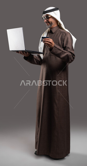 استخدام التقنيات والوسائل الحديثة والمطورة، إمكانية تسديد الرسوم والفواتير عبر الانترنت، بورتريه لرجل كبير بالسن عربي خليجي سعودي مبتسم يرتدي الثوب والغترة التقليدية يمسك بيده اللابتوب والبطاقة البنكية بإيماءات السعادة، صورة الجسم بالكامل، خلفية رمادية.