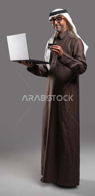 التسوق الالكتروني الآمن عبر الانترنت، النظر إلى الكاميرا بإيماءات السرور باستخدام الوسائل الحديثة، بورتريه لرجل شايب عربي خليجي سعودي مبتسم يرتدي الثوب والغترة التقليدية يمسك بيده اللابتوب وبطاقة الإئتمان البنكية، صورة الجسم بالكامل، خلفية رمادية.