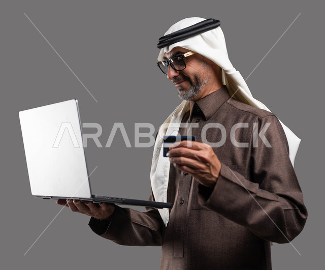 إمكانية تسديد الرسوم والفواتير عبر الانترنت، استخدام التقنيات والوسائل الحديثة والمطورة، بورتريه مقرب لرجل كبير بالسن عربي خليجي سعودي مبتسم يرتدي الثوب والغترة التقليدية يمسك بيده اللابتوب والبطاقة البنكية بإيماءات السعادة، خلفية رمادية.