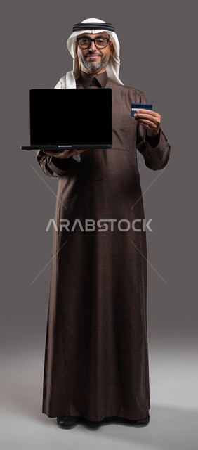 الدفع الالكتروني عبر الانترنت، النظر إلى الكاميرا بإيماءات السرور باستخدام الوسائل الحديثة، بورتريه لرجل شايب عربي خليجي سعودي مبتسم يرتدي الثوب والغترة التقليدية يمسك بيده اللابتوب بشاشة سوداء فارغة وبطاقة الإئتمان البنكية، صورة الجسم بالكامل، خلفية رمادية.