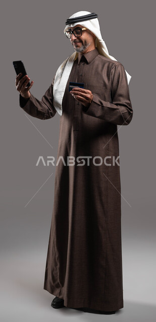 Purchasing, paying and paying bills through mobile programs and applications, portrait of an old Saudi Gulf Arab man wearing the traditional thobe and ghutra, holding a mobile phone and bank credit card in his hand with gestures of pleasure with technology, full body portrait, gray background.